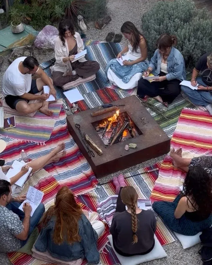 Ronda de personas sentadas sobre mantas alrededor de un fuego, escribiendo durante un encuentro al aire libre