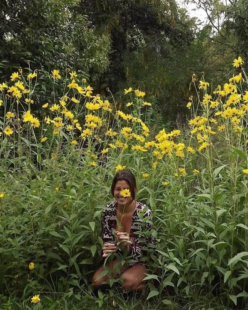 Kara Escande sonriendo entre flores amarillas, con una flor en la mano